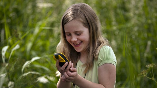 A smiling girl looks at a handheld GPS device at Wicken Fen National Nature Reserve, Cambridgeshire, against a background of tall green plants